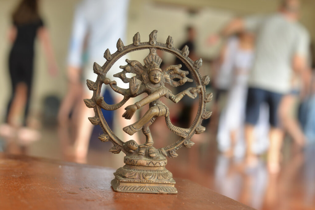 Escultura de Shiva Nataraja simbolizando o Tantra como caminho de transformação, consciência corporal e alquimia interior, com praticantes ao fundo em vivência tântrica.